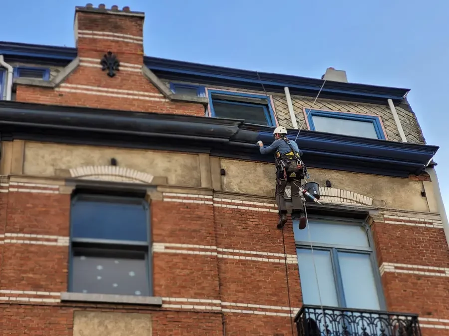 Cordiste à Bruxelles de l'entreprise Hissé ô, ponçage et peinture d'une corniche bleue en étant suspendu sur cordes. Immeuble en briques de Schaerbeek